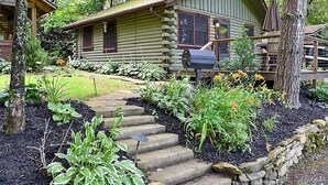 Una cabaña de troncos con una escalera de piedra que lleva a la puerta principal, rodeada de un jardín con diversas plantas y flores.