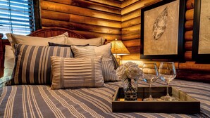 Una habitación acogedora con cama, almohadas, una bandeja con un florero y copas de vino, y un cuadro de una hoja en la pared.