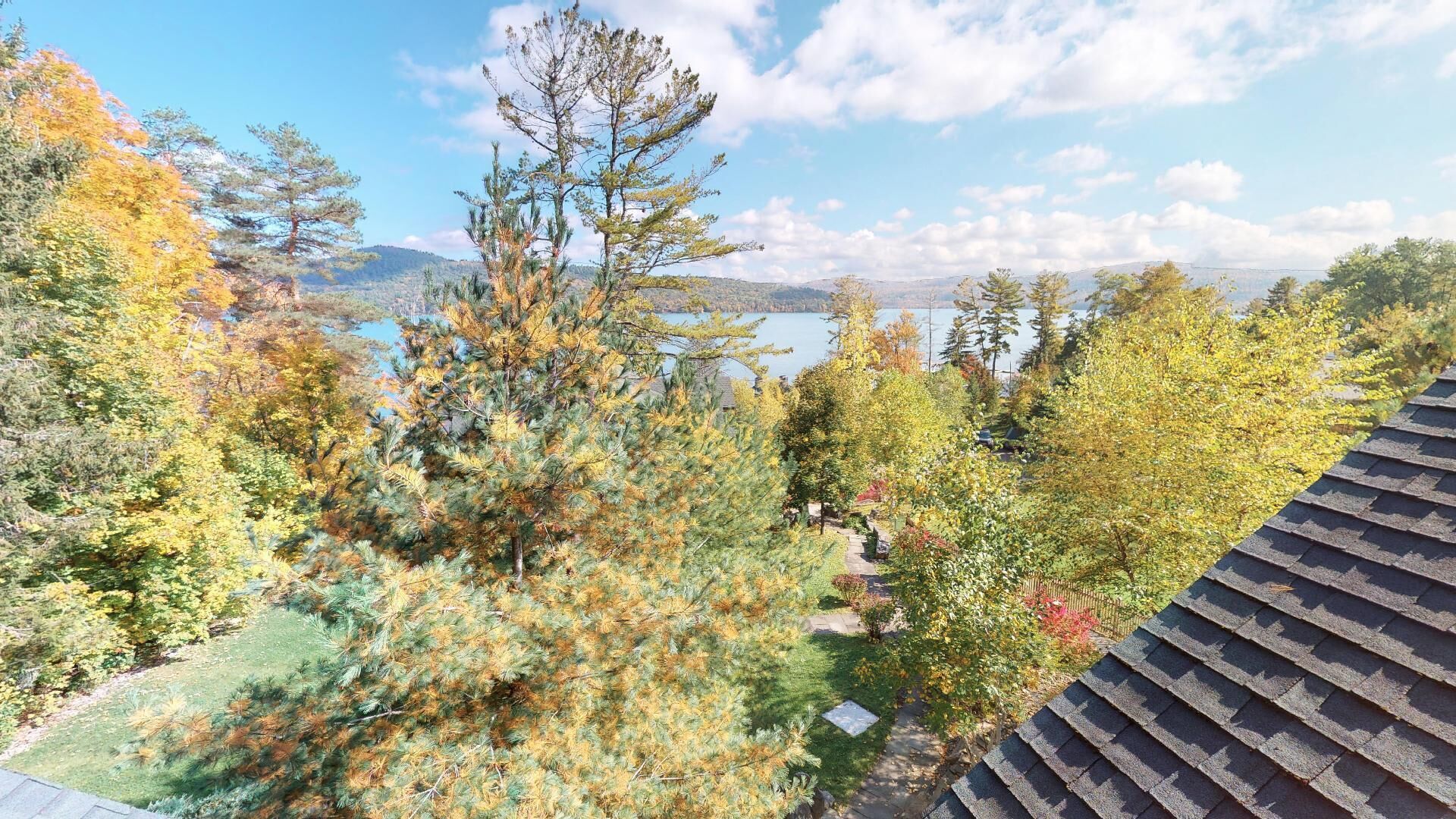 Zona residencial con vistas a un lago y montañas. Hay árboles con follaje otoñal, un sendero pavimentado y el borde de un tejado.