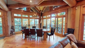 A spacious room with a wooden dining table and chairs, large windows offering views of trees, and a wooden ceiling with exposed beams.