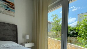 A hotel room with a large bed, a nightstand, a lamp, and a view of greenery and buildings through a large window. - Apt Tina, Modern, 2 bedrooms, Outdoor Space & BBQ, close to beach (Mastrinka)