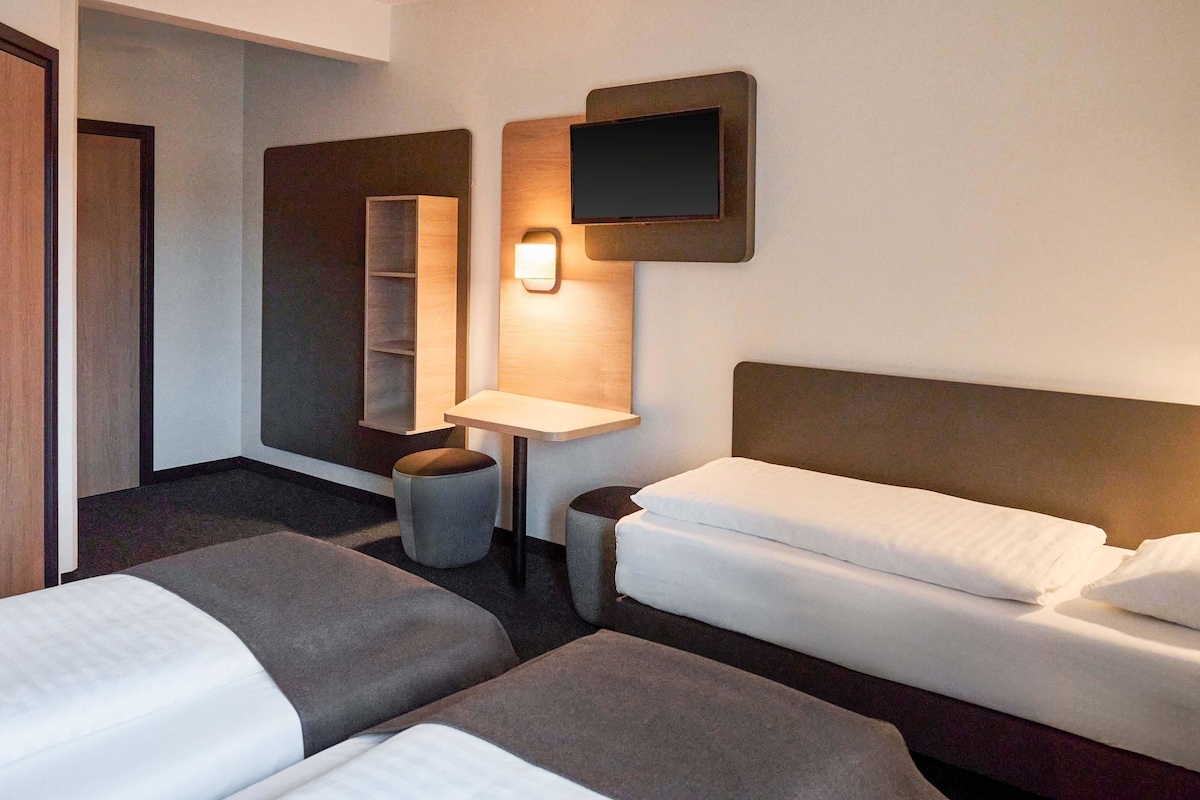 A hotel room with two beds, a small table, a chair, a TV, and a wall-mounted shelf.