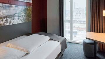 Habitación de hotel con cama, una mesita, ventana con vista a la ciudad y un cuadro en la pared.
