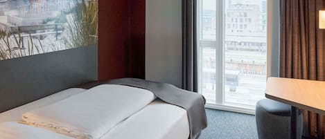 Habitación de hotel con cama, una mesita, ventana con vista a la ciudad y un cuadro en la pared.