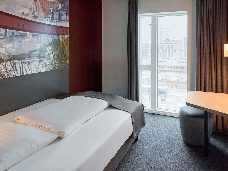 A hotel room with a bed, a small table, a window with a city view, and a painting on the wall.