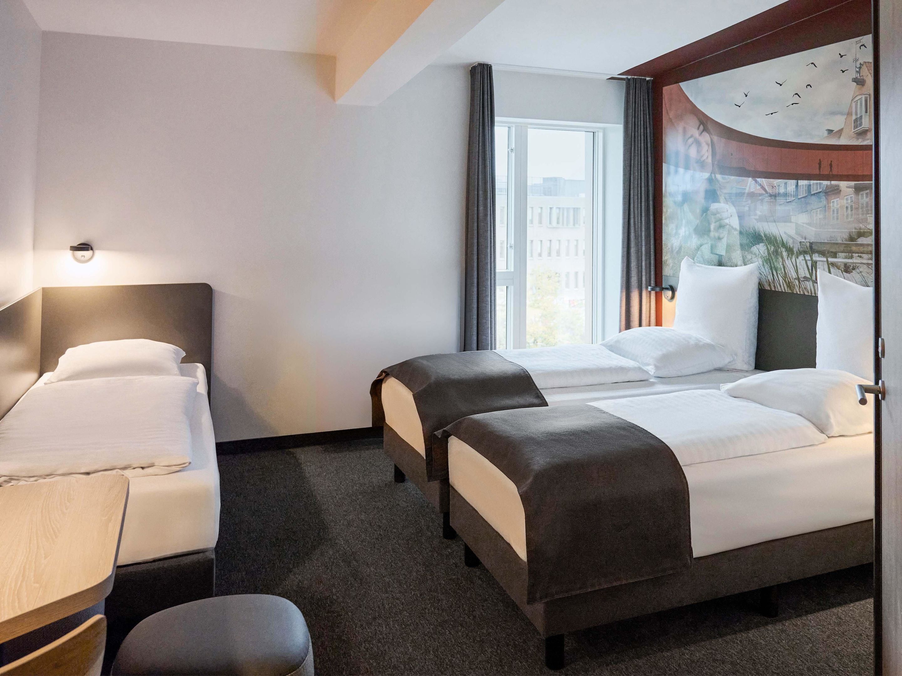 A hotel room with two beds, a desk, and a large window with a view of buildings and birds.