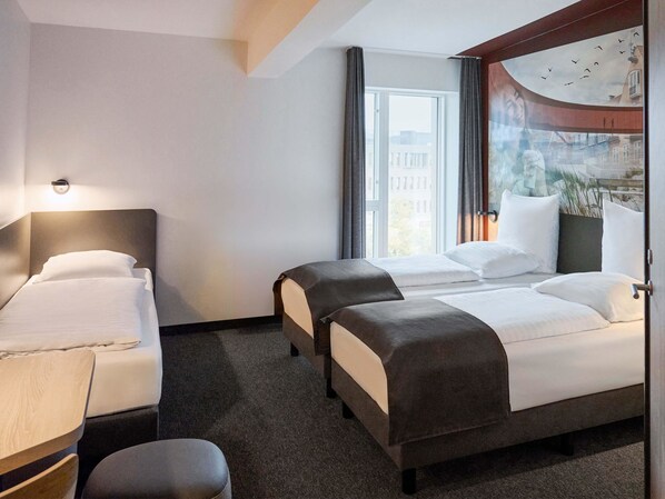 A hotel room with two beds, a desk, and a large window with a view of buildings and birds. - B&B Hotel Vejle (Vejle)