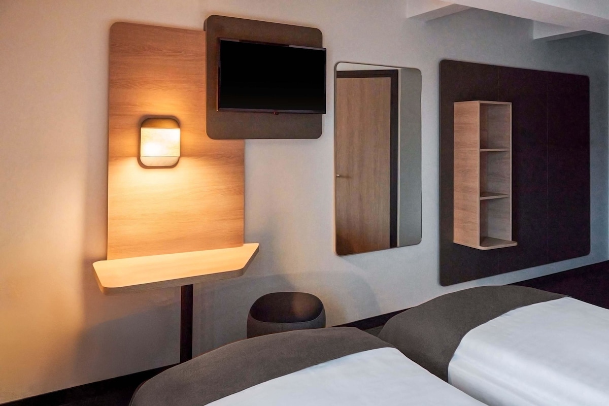 A hotel room with two beds, a TV mounted on a wooden panel, a mirror, and a shelf with a light.