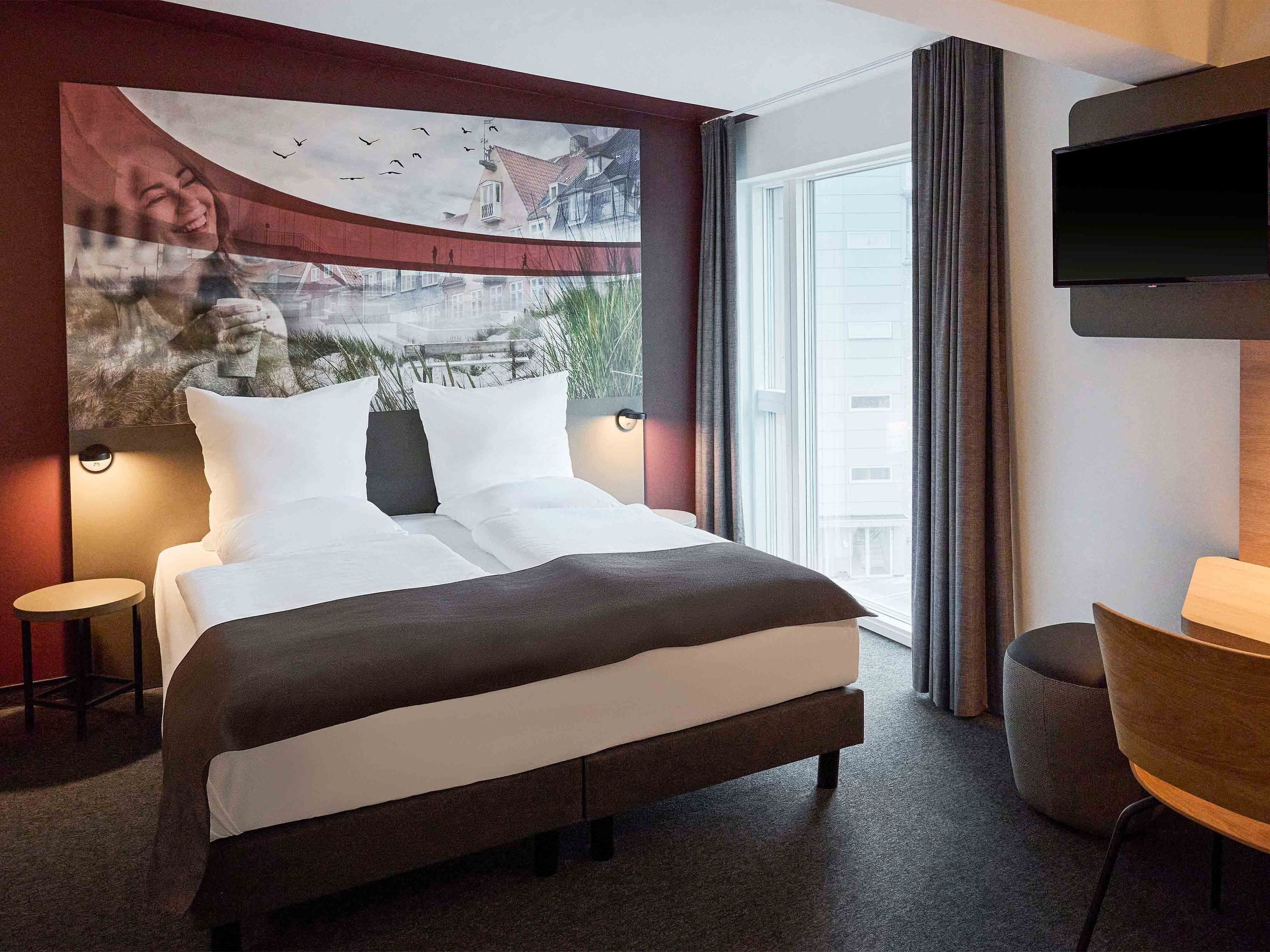 A hotel room with a large bed, a small table, a chair, a TV, and a window with curtains.
