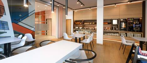A modern cafe with white tables and chairs, a bar area with a counter and shelves, and a staircase in the background.
