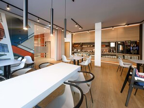 Un café moderne avec des tables et des chaises blanches, un coin bar doté d’un comptoir et d’étagères, et un escalier en arrière-plan.