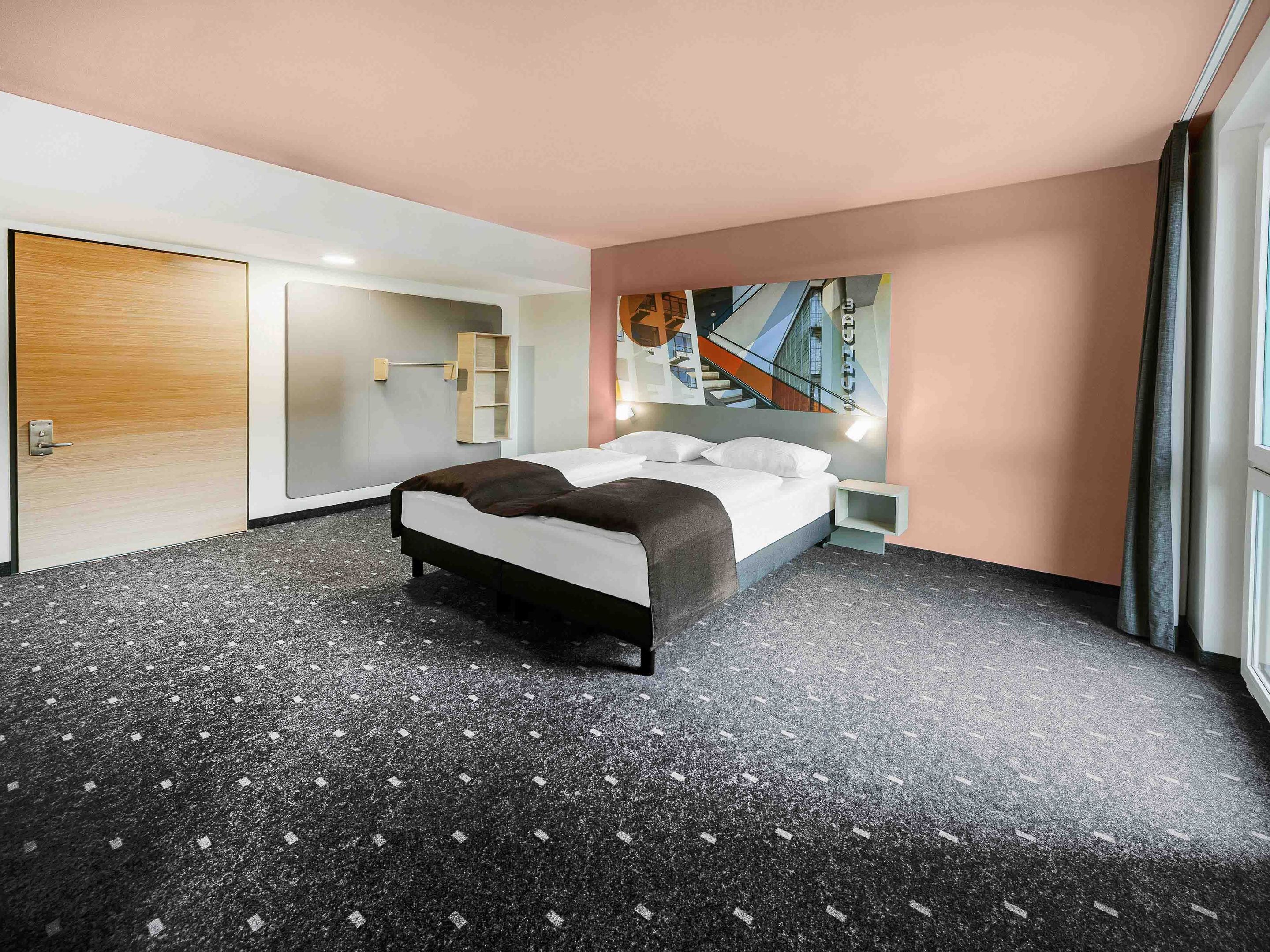 A modern hotel room with a large bed, a small bedside table, a built-in shelf, and a painting on the wall.