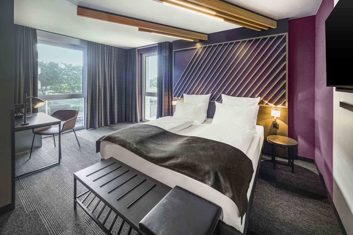 Una habitación de hotel moderna con una cama grande, un escritorio y una silla. Cuenta con una ventana con cortinas, un televisor montado en la pared y una mesita de noche con una lámpara.