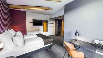 Ett modernt hotellrum med en soffa, ett skrivbord, en stol, en platt-TV och en bokhylla.