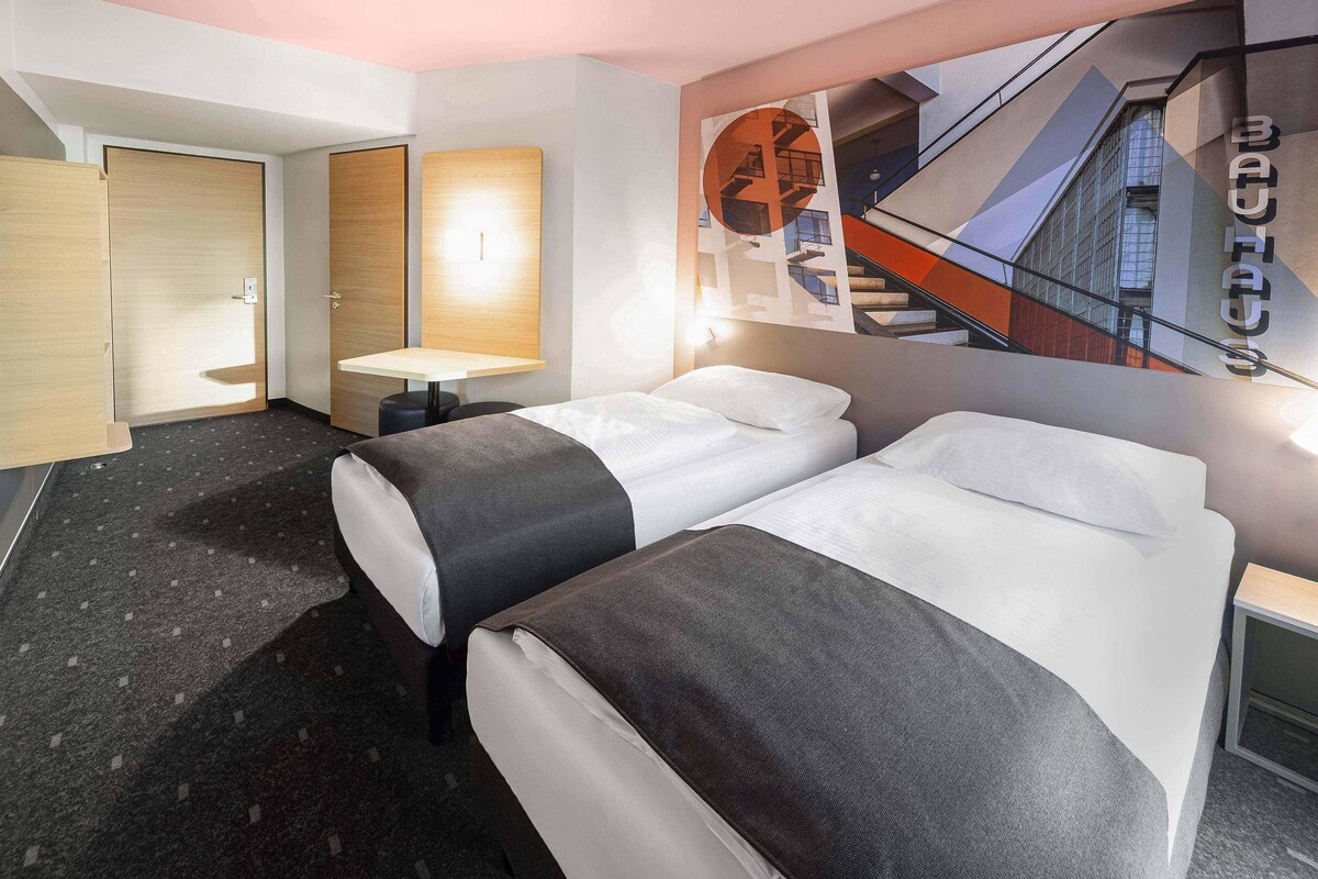 Une chambre d’hôtel avec deux lits, une petite table, une lampe fixée au mur et une grande fenêtre donnant sur un immeuble.