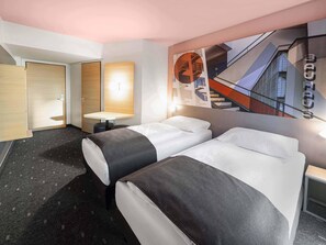 Een hotelkamer met twee bedden, een klein tafeltje, een wandlamp en een groot raam met uitzicht op een gebouw.