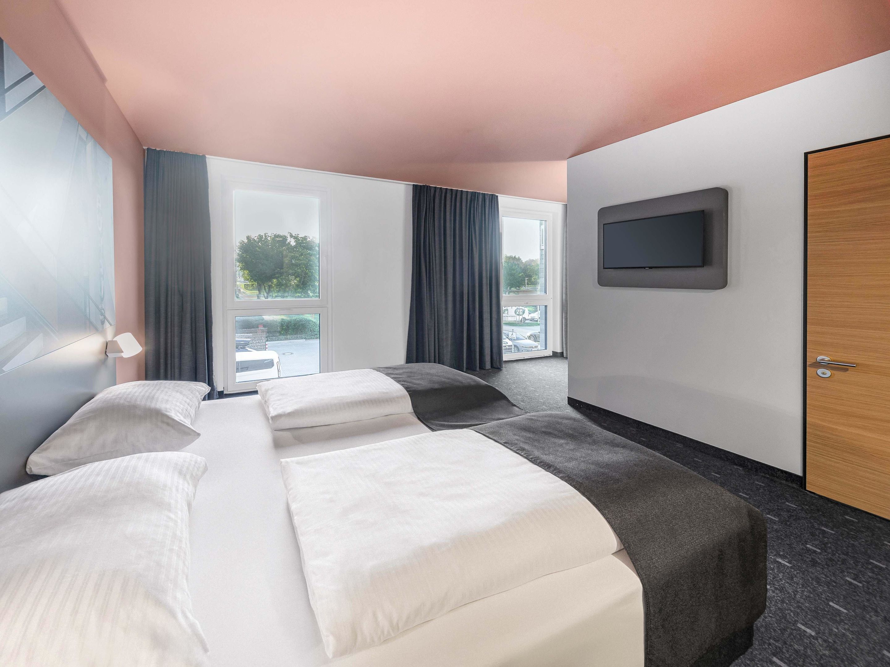 A hotel room with a bed, a TV mounted on the wall, and a large window with curtains.