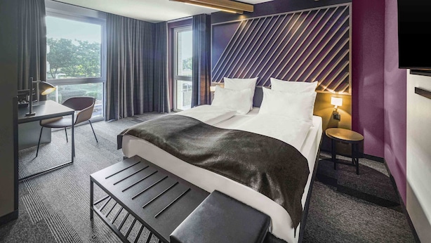 Een moderne hotelkamer met een groot bed, een bureau en een stoel. Er is een raam met gordijnen, een wandtelevisie en een tafeltje met een lamp.