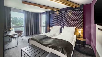 Ett modernt hotellrum med en stor säng, ett skrivbord och en stol. Det finns ett fönster med gardiner, en väggmonterad TV och ett litet bord med en lampa.