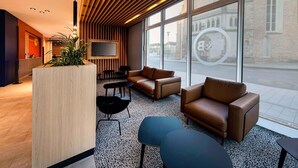 Een moderne lobby met een receptiebalie, een zithoek en grote ramen met uitzicht op de straat.