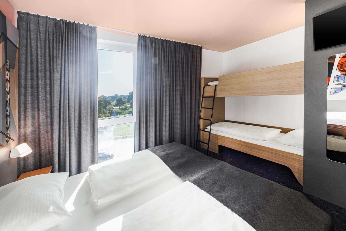 Une chambre d’hôtel avec un lit superposé, un bureau, une chaise et une grande fenêtre avec des rideaux.