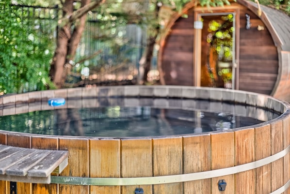 Un jacuzzi con revestimiento de madera, rodeado de follaje y una estructura de madera al fondo.