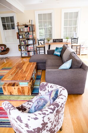 Una sala de estar con un sofá modular, un sillón estampado, una mesa de centro de madera, una estantería y una zona de comedor con mesa y sillas.