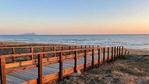 Ein hölzerner Steg führt zu einem Strand mit freiem Blick auf das Meer und ein fernes Gebirge.