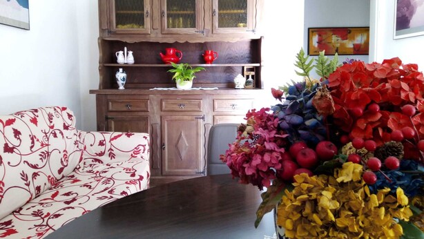 Una sala de estar con un sofá floral, un mueble de madera con puertas de vidrio y un jarrón grande de flores.