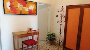 Una habitación con un escritorio de madera, una silla roja, una planta en maceta, un soporte metálico y un armario grande.