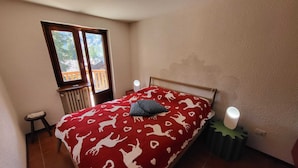 A bedroom with a bed, a window, a radiator, a small table, and a bedside lamp. - Mont Blanc Apt- Bluchalet  (Courmayeur)