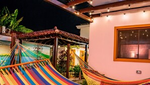 Een hangmat onder een houten pergola met lichtslingers, voor een witte muur met een raam.