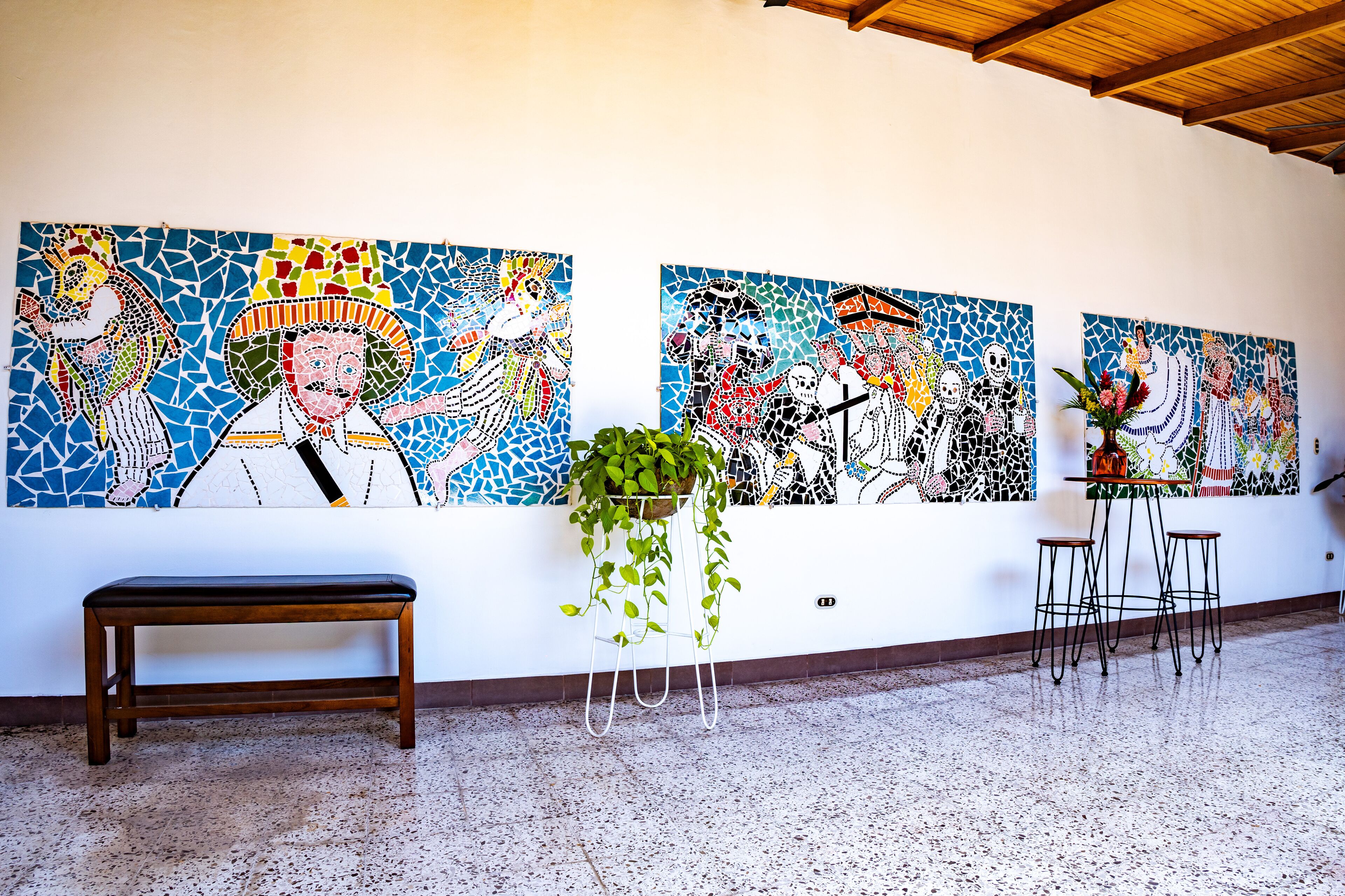 Una habitación con suelo de baldosas, un banco de madera, una planta en maceta y una pared con arte en mosaico.