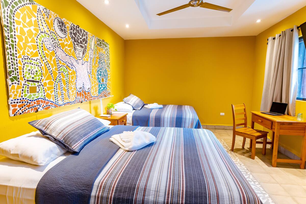 Une chambre d’hôtel avec deux lits, un bureau, une chaise, un ventilateur de plafond et une grande œuvre d’art en mosaïque au mur.