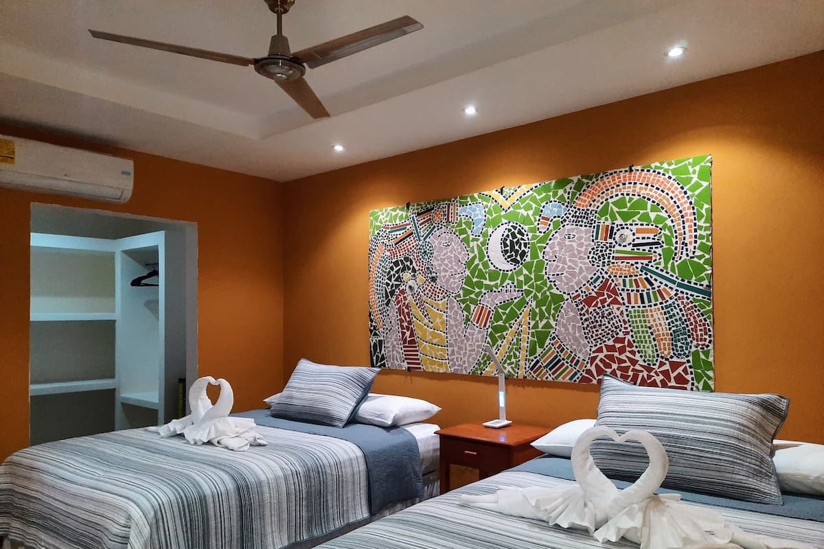 Une chambre d’hôtel avec deux lits, un ventilateur de plafond, un mur orné d’une mosaïque colorée et une armoire.
