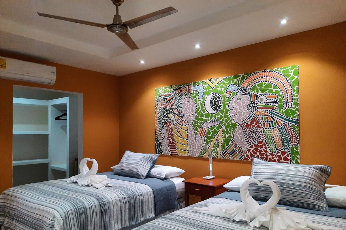Une chambre d’hôtel avec deux lits, un ventilateur de plafond, un mur orné d’une mosaïque colorée et une armoire.