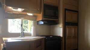 A compact kitchen with a microwave, stove, and sink, featuring wooden cabinets and a window providing natural light. - Misty Haven Farm (Ladysmith)