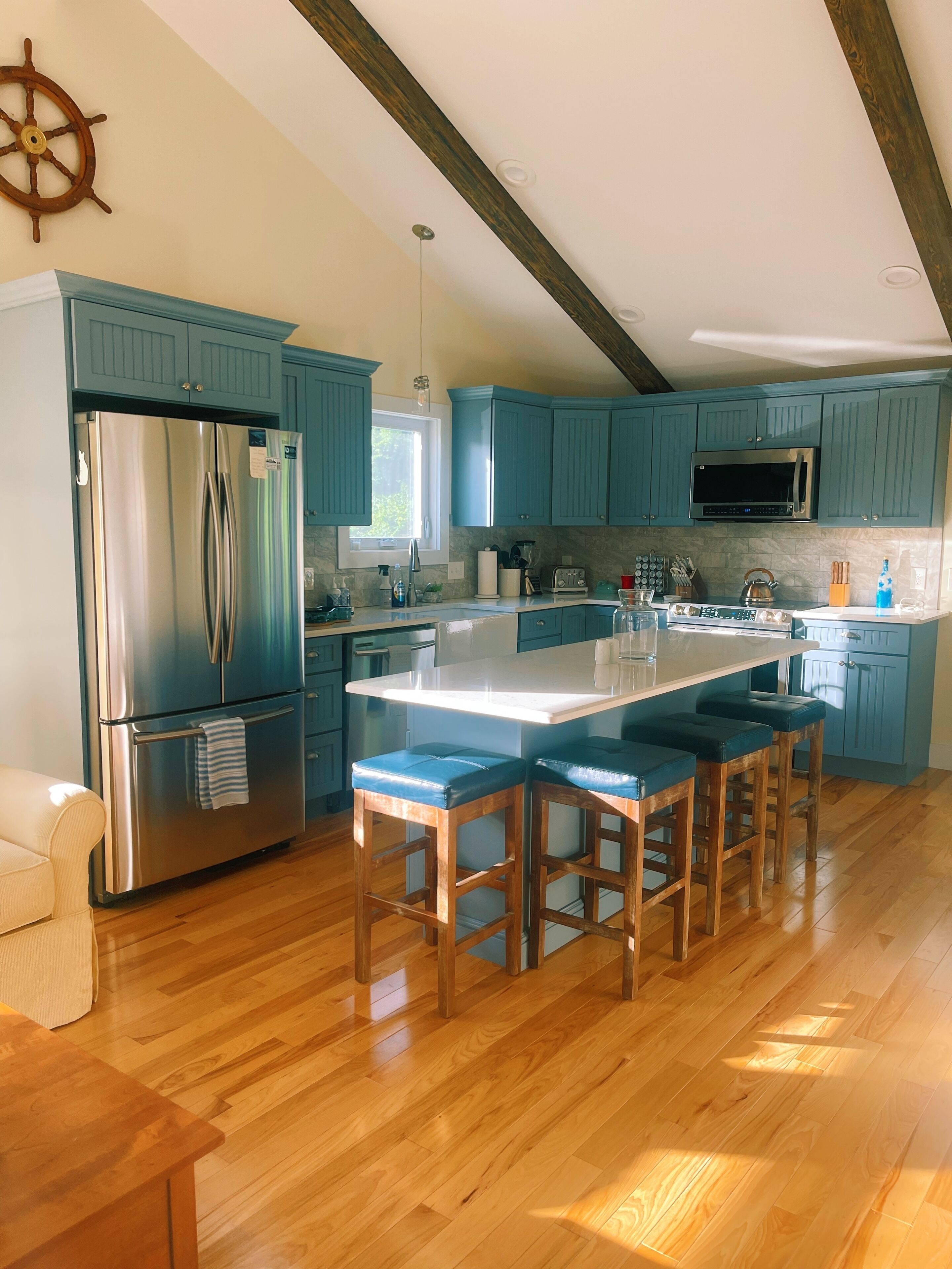 Une cuisine moderne avec des meubles de couleur turquoise, un réfrigérateur en acier inoxydable, un îlot central doté de tabourets de bar et des poutres en bois au plafond.