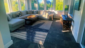 Una sala de estar moderna con un sofá seccional, una mesa de centro y una alfombra. Amplias ventanas ofrecen luz natural y vistas a la vegetación exterior.