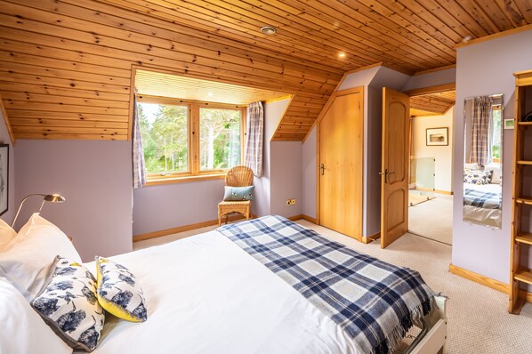 Un dormitorio con techo de madera, una cama grande con manta a cuadros, una silla de mimbre y una ventana con cortinas.