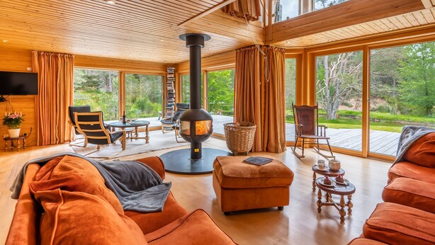 Een gezellig interieur van een blokhut met een houtkachel, grote ramen met uitzicht op het bos, en een comfortabele zithoek met een bank en fauteuils.