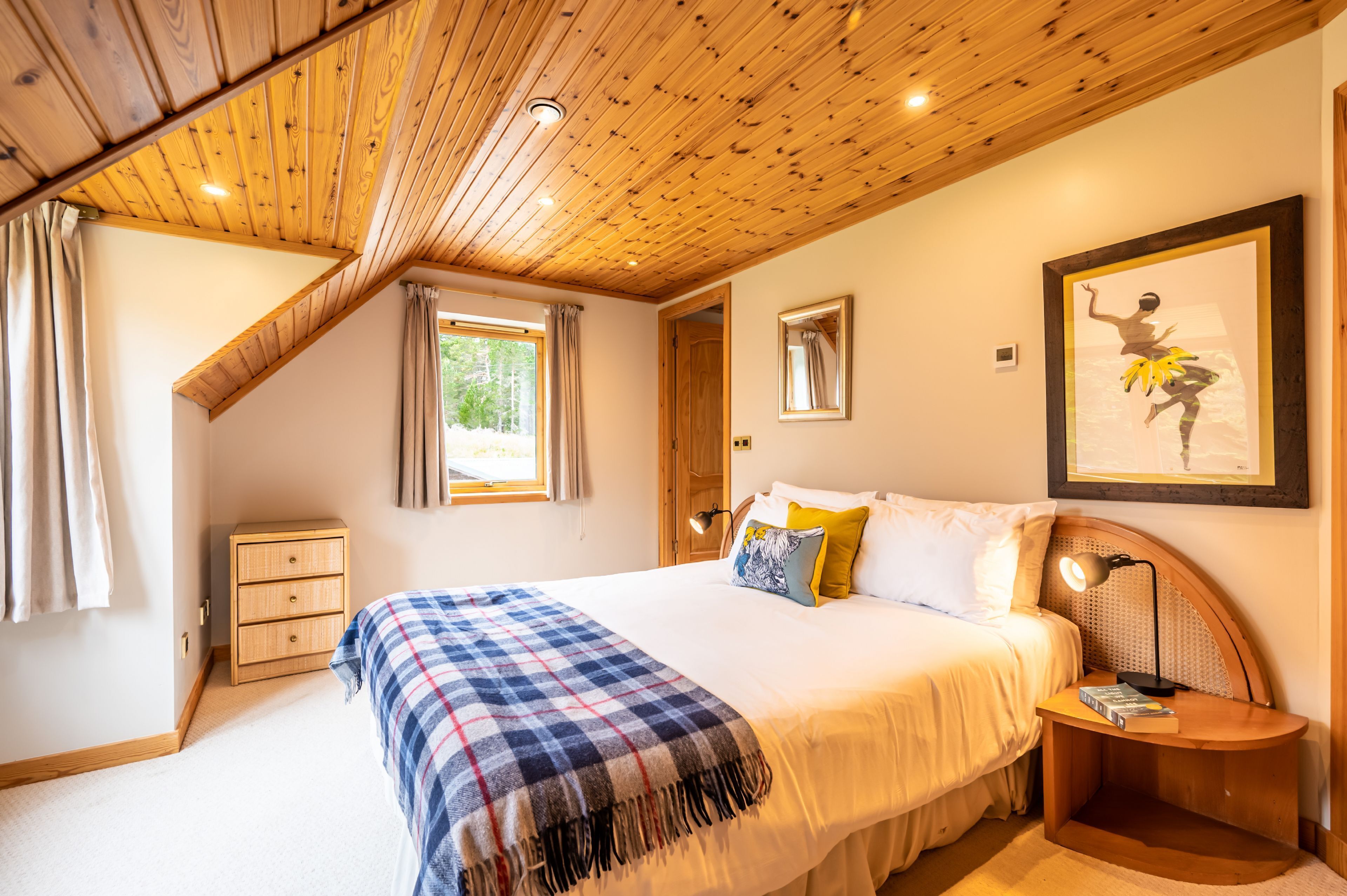 A bedroom with a wooden ceiling, a bed with a plaid blanket, a nightstand with a lamp, and a framed picture on the wall.