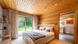 Een slaapkamer met een groot bed, een houten plafond, een schuifdeur naar een tuin en een kleine badkamer met een handdoekhouder.