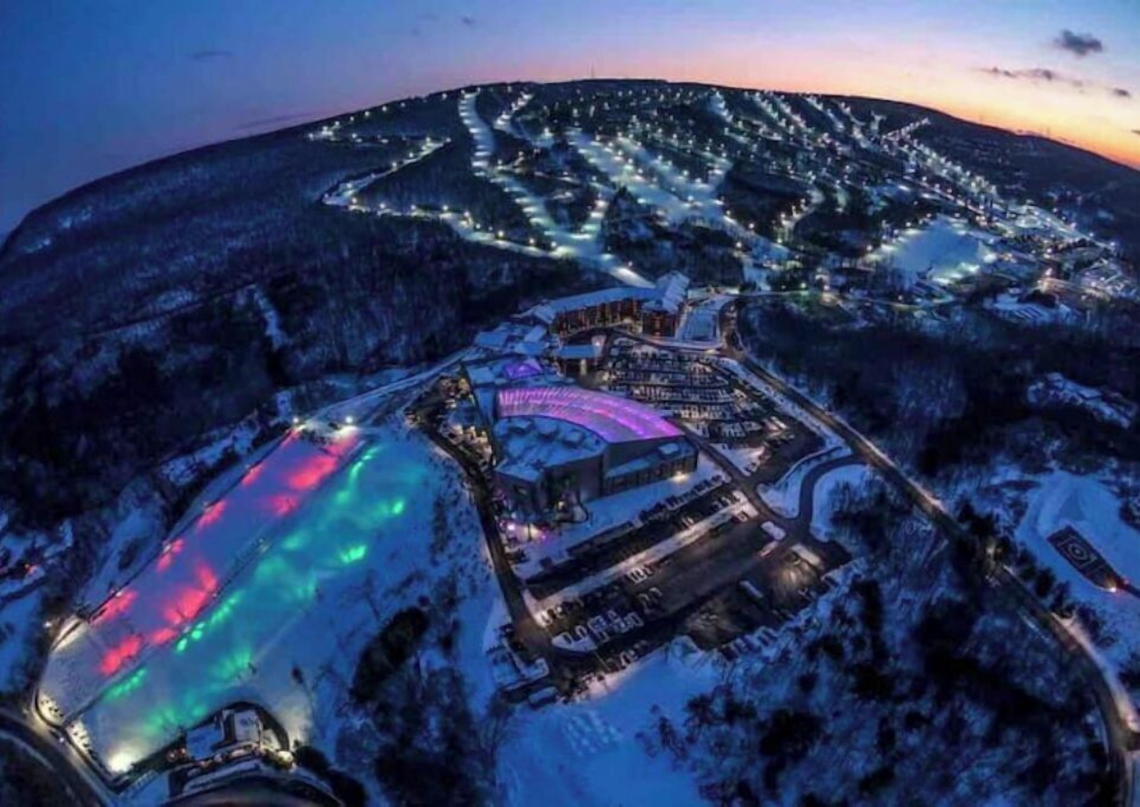 An aerial view of a snow-covered resort with illuminated pathways and a central building with a colorful roof.