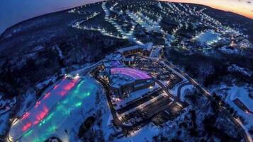 An aerial view of a snow-covered resort with illuminated pathways and a central building with a colorful roof.