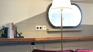 A room with a red sofa, patterned pillows, a floor lamp, a round window, and a shelf with books and decorative items. - Atypical accommodation on a barge (Ronquière)