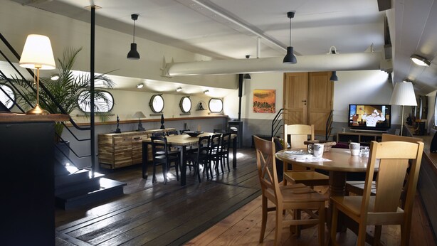 A dining area with wooden tables and chairs, a kitchen in the background, and a television on the right. - Atypical accommodation on a barge (Ronquière)