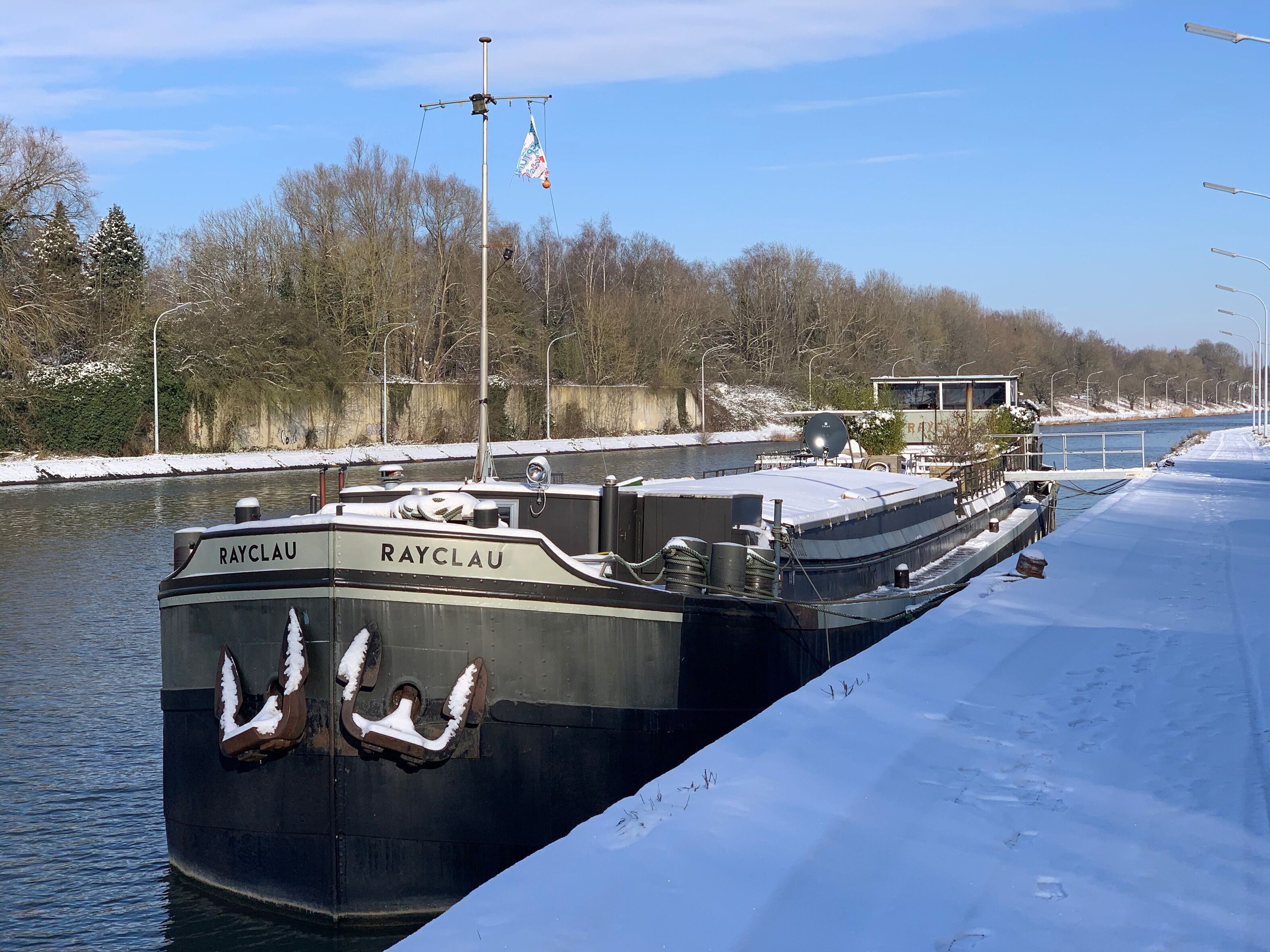 A boat named 'RAYCLAU' docked at a pier with a snowy landscape and trees in the background.