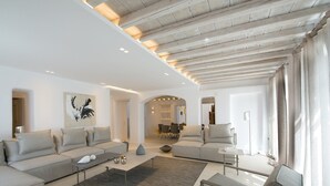 A modern living room with a sectional sofa, a coffee table, and a side table. There is a large window with curtains, a ceiling with exposed beams, and a painting on the wall. - ISholidays Mykonos Villa Zircon (Tourlos)
