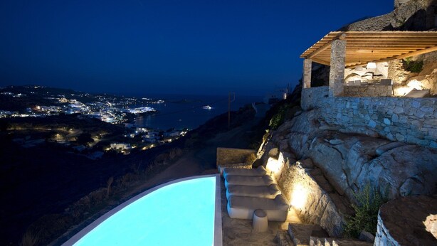Un bâtiment en pierre doté d’une piscine et d’un espace extérieur aménagé, offrant une vue sur une ville côtière la nuit.